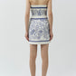 A woman stands with her back to the camera, wearing the Simkhai Darbie Linen Mini Dress in a blue and white seashell print with a wrap silhouette, paired with white sandals, set against a plain background.