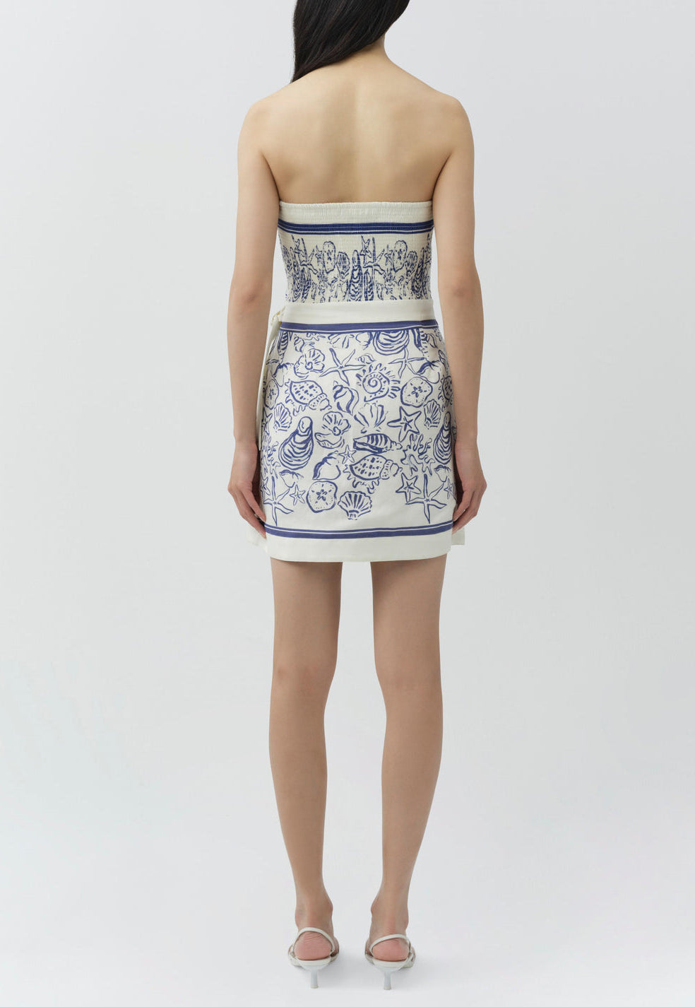 A woman stands with her back to the camera, wearing the Simkhai Darbie Linen Mini Dress in a blue and white seashell print with a wrap silhouette, paired with white sandals, set against a plain background.