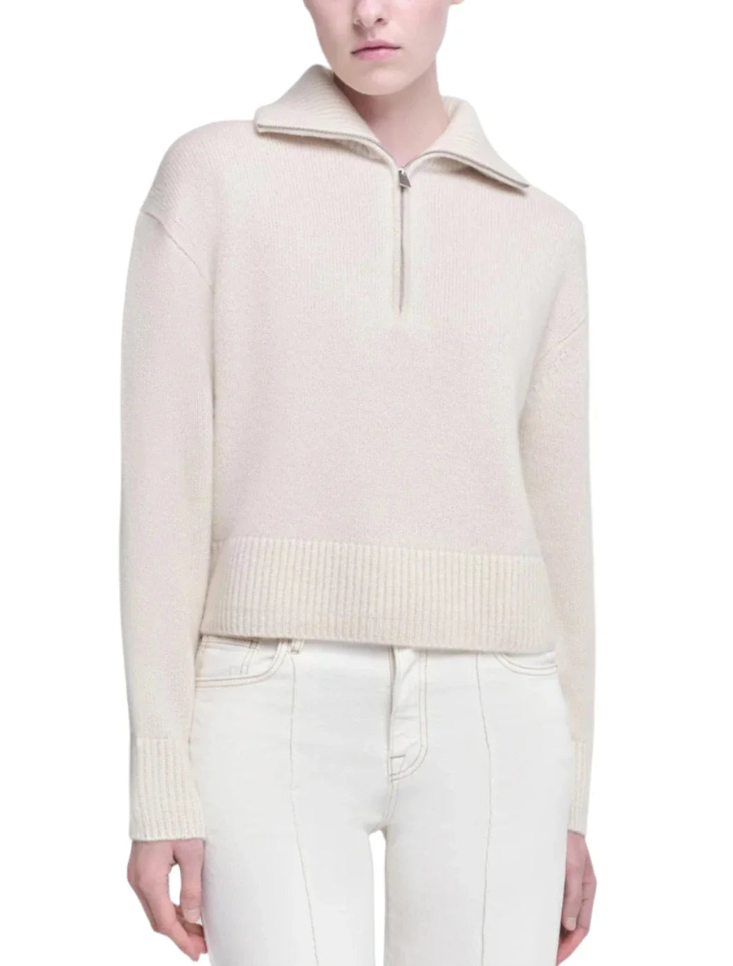 A person wears a cream-colored, long-sleeve sweater with a half-zip closure and white pants. The luxurious Simkhai Andes Half Zip Pullover is crafted from 100% cashmere, adding both style and comfort to the ensemble.