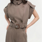 A person wears the Simkhai Axel Top, a taupe short-sleeve piece with a double-breasted closure, belted waist, and utilitarian notch collar, standing against a plain light background.