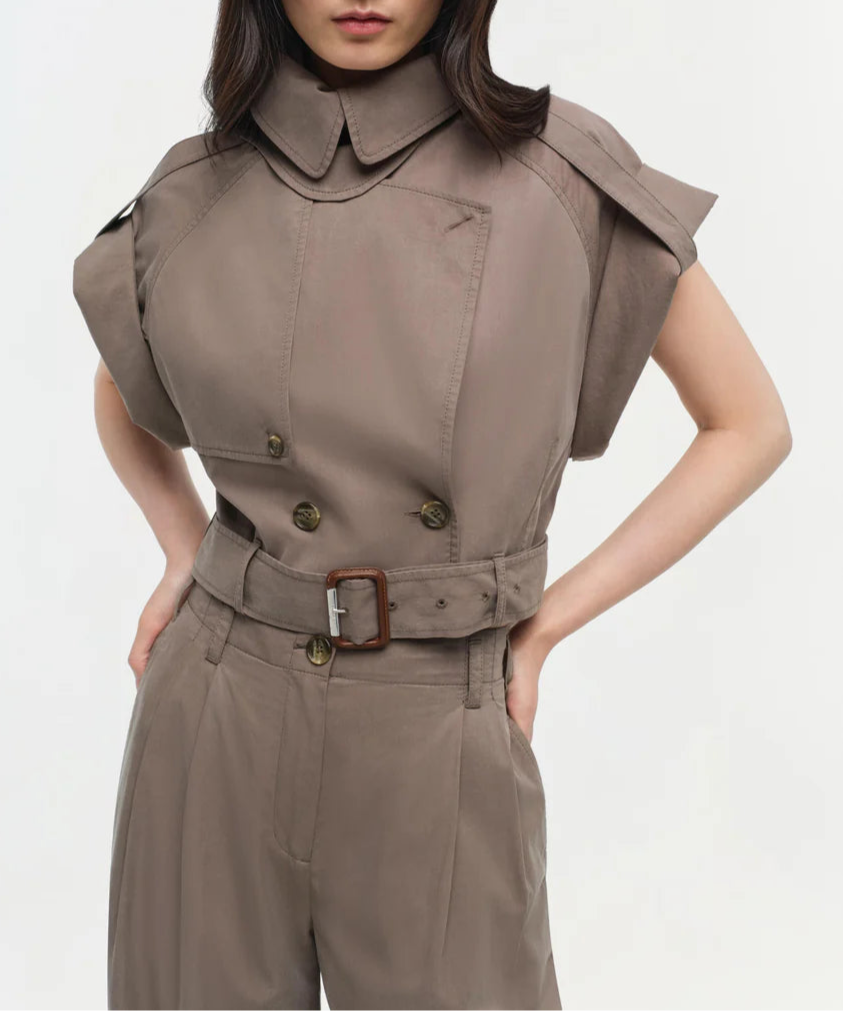 A person wears the Simkhai Axel Top, a taupe short-sleeve piece with a double-breasted closure, belted waist, and utilitarian notch collar, standing against a plain light background.
