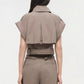A woman is seen from the back wearing the Simkhai Axel Top, a taupe, short-sleeve cropped jacket with a utilitarian notch collar and vented back detail, paired with matching high-waisted pants.