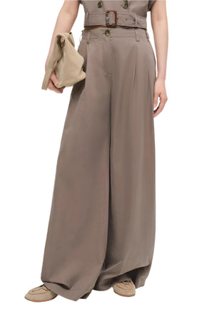 A person wears the Simkhai Haydin Wide Leg Pant—high-waisted, ultra-wide linen pants with a button front, side pockets, and matching belt—paired with a beige clutch and beige loafers.