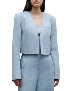 A woman models the TWP Drink At Costes set: a light blue cropped blazer with shoulder pads, worn over a matching top and high-waisted pants, hands in her pockets.