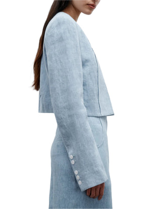A woman wears TWP’s “Drink At Costes,” a light blue long-sleeved cropped blazer with shoulder pads, four white cuff buttons, and a matching high-waisted skirt.