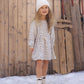 A young child with long curly hair wearing a white beanie and the Rylee & Cru Girls’ Tate Dress—an animal print dress with a gathered waist and adjustable drawstring—paired with boots, stands on snow in front of a wooden fence and vintage sleds.