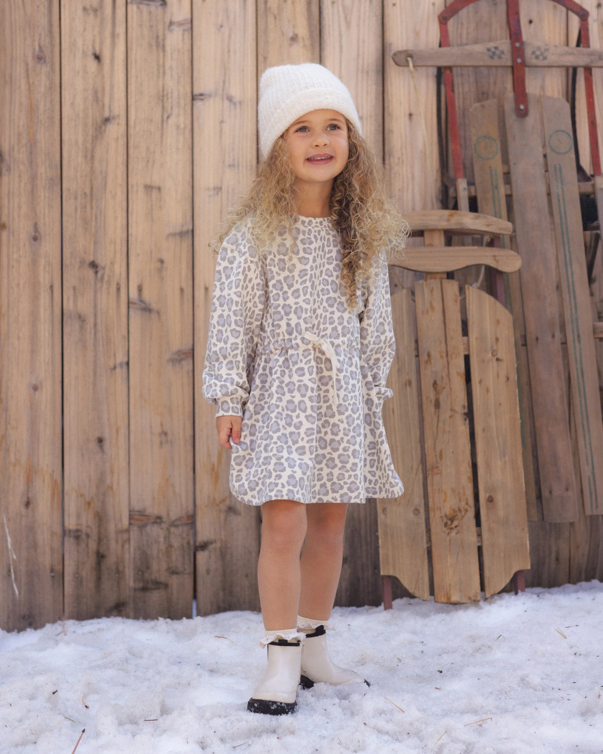 A young child with long curly hair wearing a white beanie and the Rylee & Cru Girls’ Tate Dress—an animal print dress with a gathered waist and adjustable drawstring—paired with boots, stands on snow in front of a wooden fence and vintage sleds.
