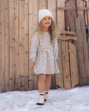 A young child with long curly hair wearing a white beanie and the Rylee & Cru Girls’ Tate Dress—an animal print dress with a gathered waist and adjustable drawstring—paired with boots, stands on snow in front of a wooden fence and vintage sleds.