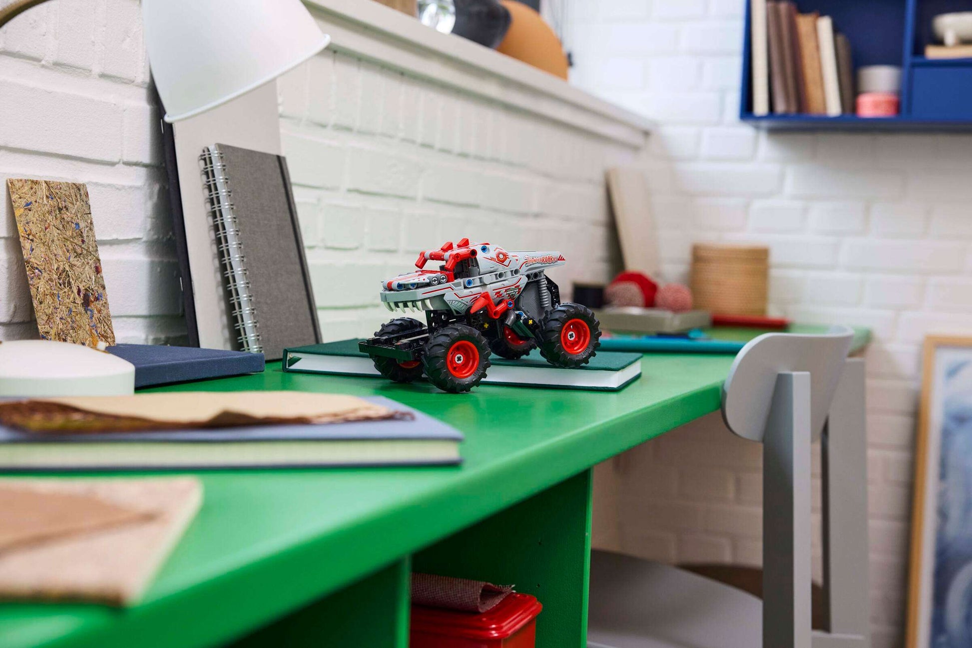 The LEGO® Technic™ Monster Jam™ ThunderROARus™ Pull-Back toy by Legos - Toyhouse is placed atop a stack of notebooks on a green desk in a study area filled with books, a lamp, and stationery items.