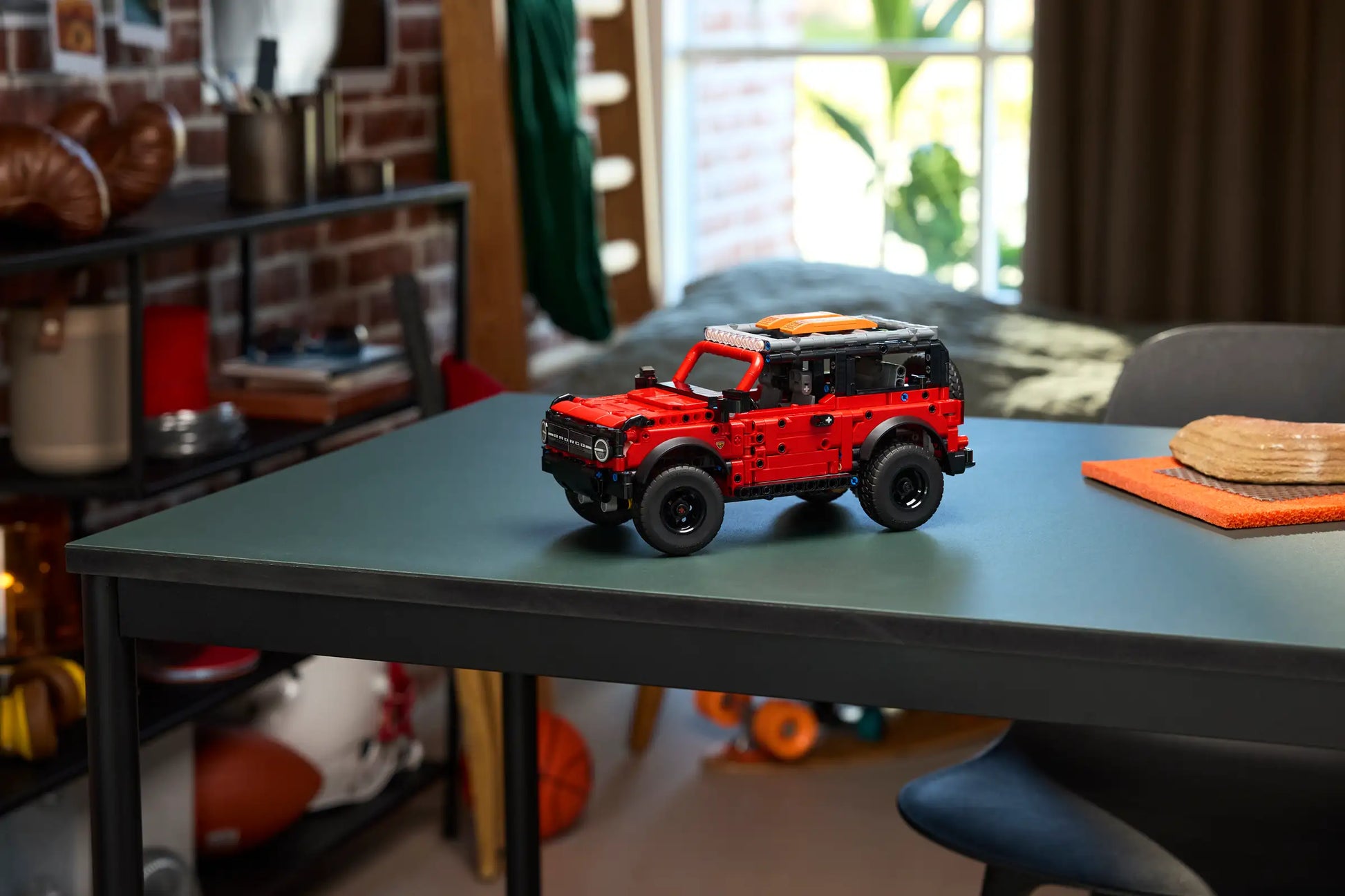 A LEGO® Technic™ Ford Bronco® SUV by Legos - Toyhouse is displayed on a dark green table in a room filled with shelves, sports equipment, and natural light.