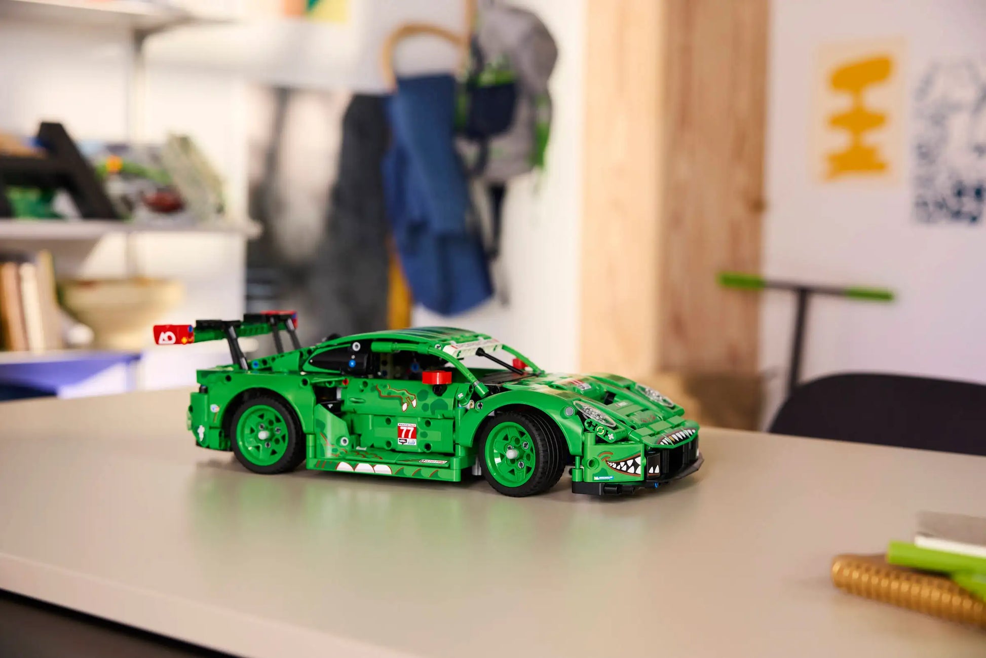 The LEGO® Technic™ Porsche 911 GT3 R REXY AO Racing Car by Legos - Toyhouse, complete with decals, is shown on a light table in a room featuring shelves, coats, and wall decor in the background.