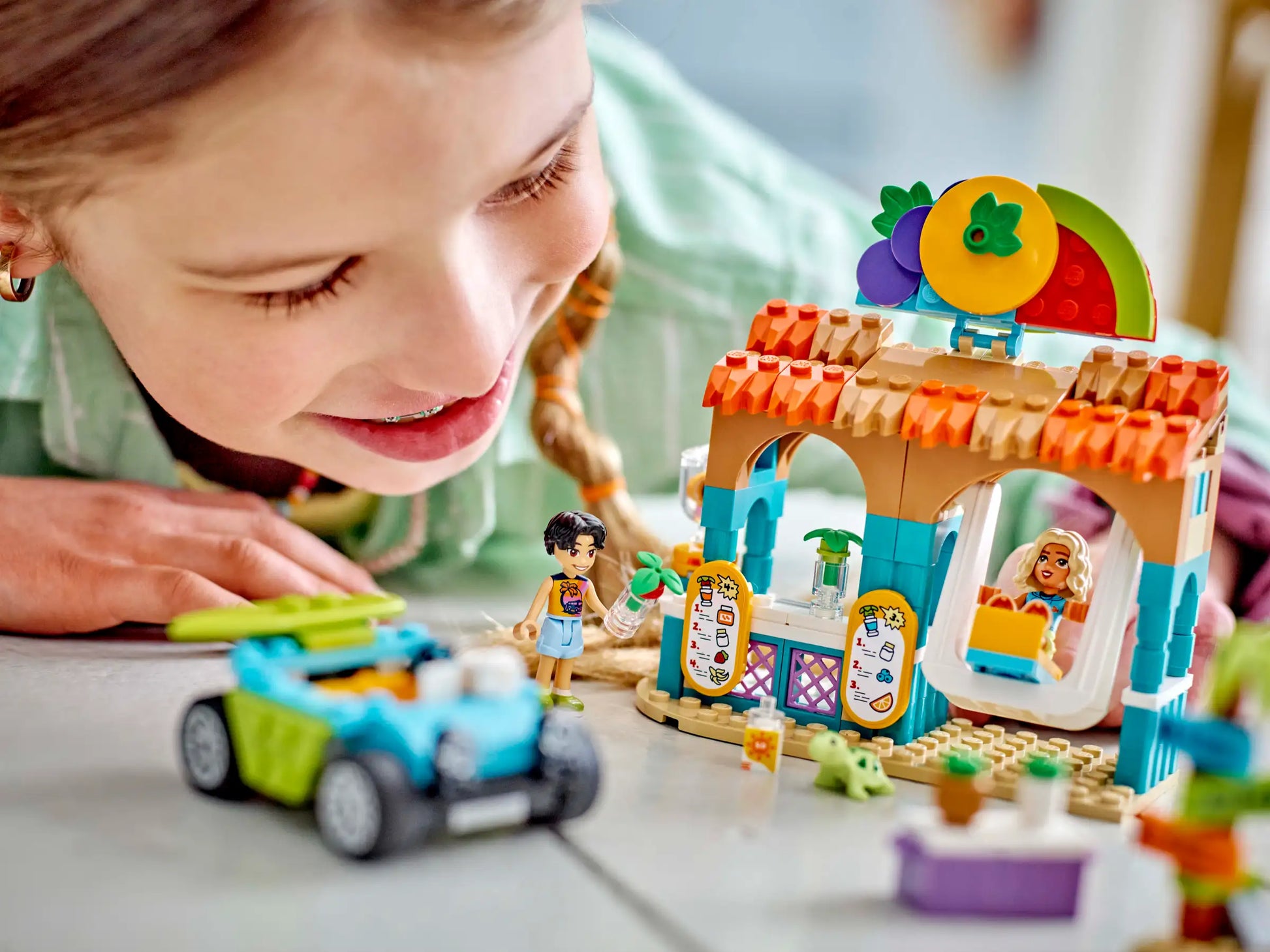 A child joyfully plays on the floor with a vibrant toy set, featuring a small car and market stall embellished with fruit decorations, reminiscent of a LEGO® Friends Beach Smoothie Stand from Legos - Toyhouse.