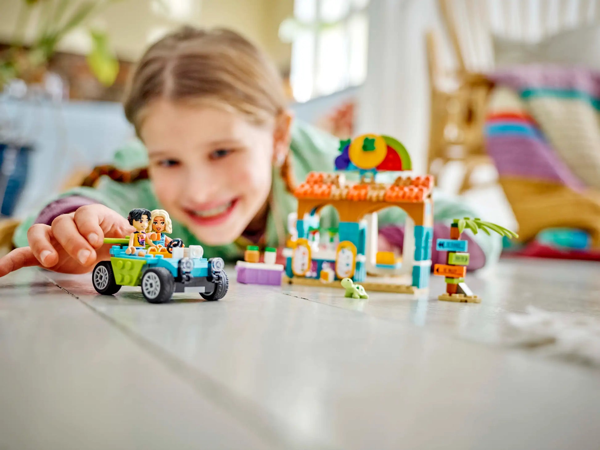 A child is deeply engaged on the floor, creatively building with toy blocks and zooming a small car around their imaginative landscape, reminiscent of a LEGO Friends adventure. Nearby, they pretend to run a beach smoothie stand using their LEGO® Friends Beach Smoothie Stand from Legos - Toyhouse playset, blending fun and creativity seamlessly.