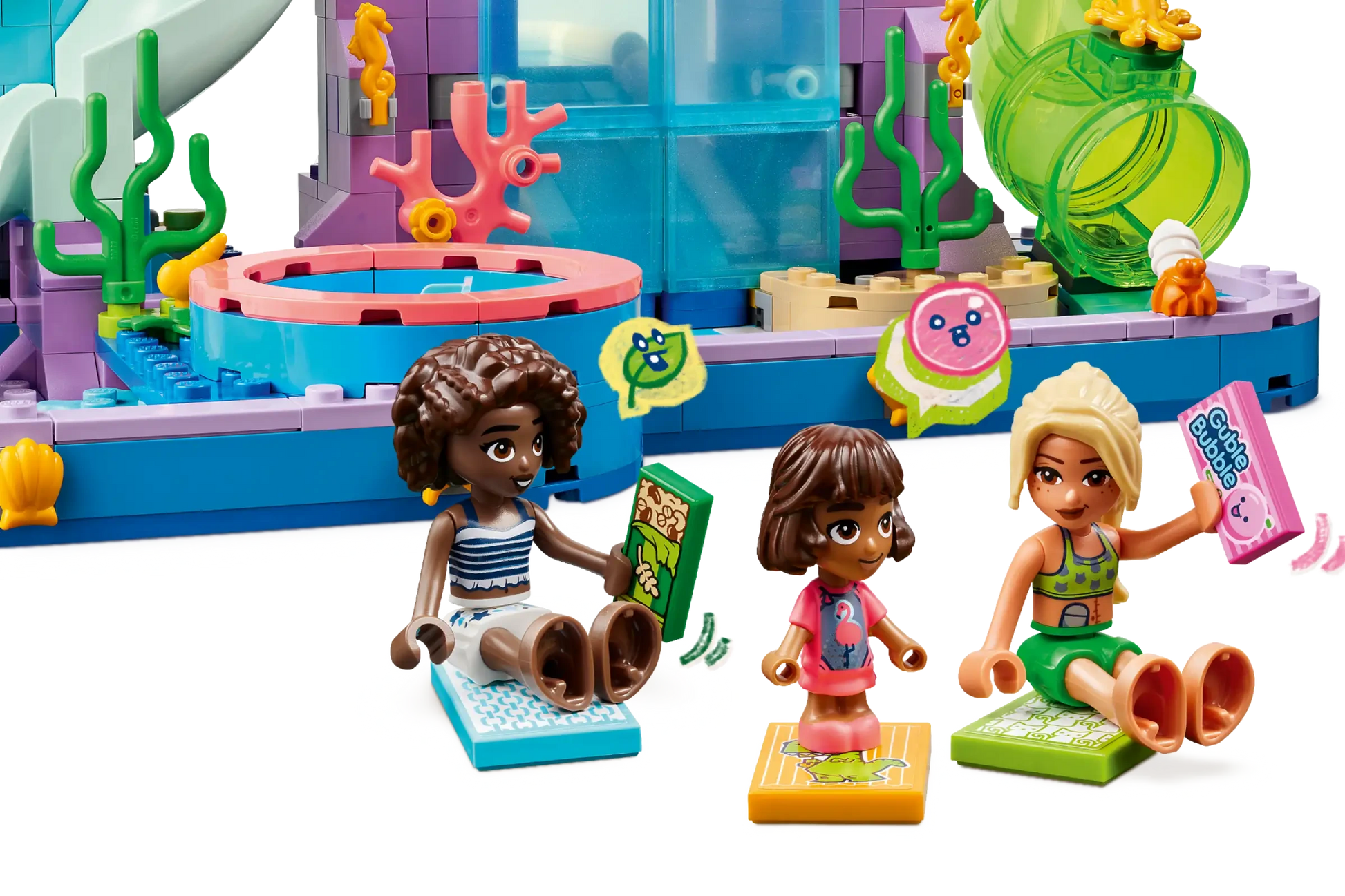 Three toy figures sit on mats with books and speech bubbles in front of the LEGO® Friends Heartlake City Water Park by Legos - Toyhouse, featuring plants and coral—perfect for kids’ imaginative role play.