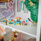 A white shelf displays colorful toy figurines, art supplies, a green textured letter "C," pastel rainbow decorations, and the Legos - Toyhouse LEGO® Friends Costume Party with Unicorn & Fairy kit for endless pretend play in a child’s room.