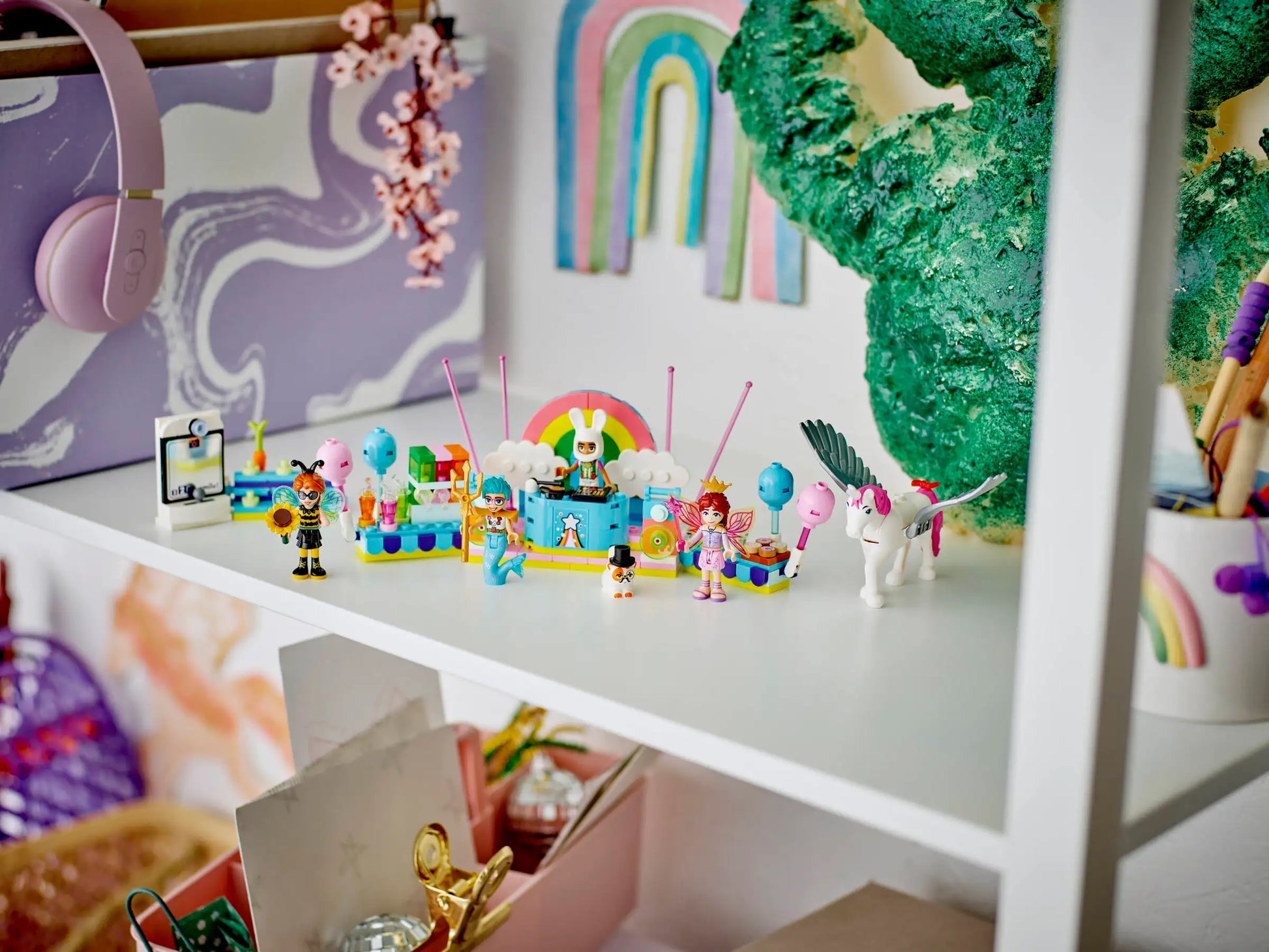 A white shelf displays colorful toy figurines, art supplies, a green textured letter "C," pastel rainbow decorations, and the Legos - Toyhouse LEGO® Friends Costume Party with Unicorn & Fairy kit for endless pretend play in a child’s room.