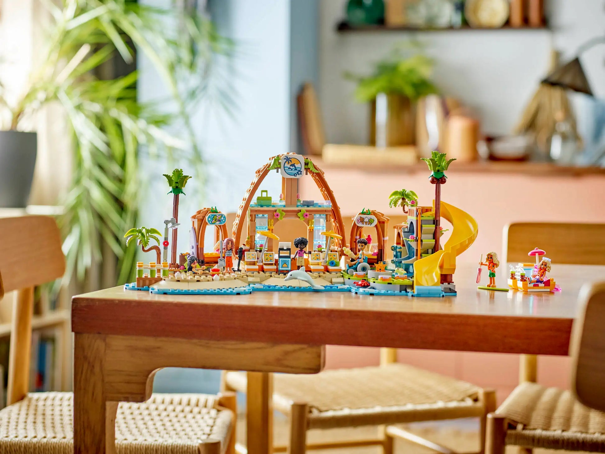 The LEGO® Friends Family Vacation Beach Resort by Legos - Toyhouse is built on a wooden dining table in a modern, bright room with wooden chairs and indoor plants.