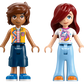 Two LEGO mini-dolls stand side by side; one with short brown hair and a patterned vest, the other with long red hair and a unicorn t-shirt—ideal for fans of unicorns and the Legos - Toyhouse LEGO® Friends Unicorn Cake Delivery Car set.