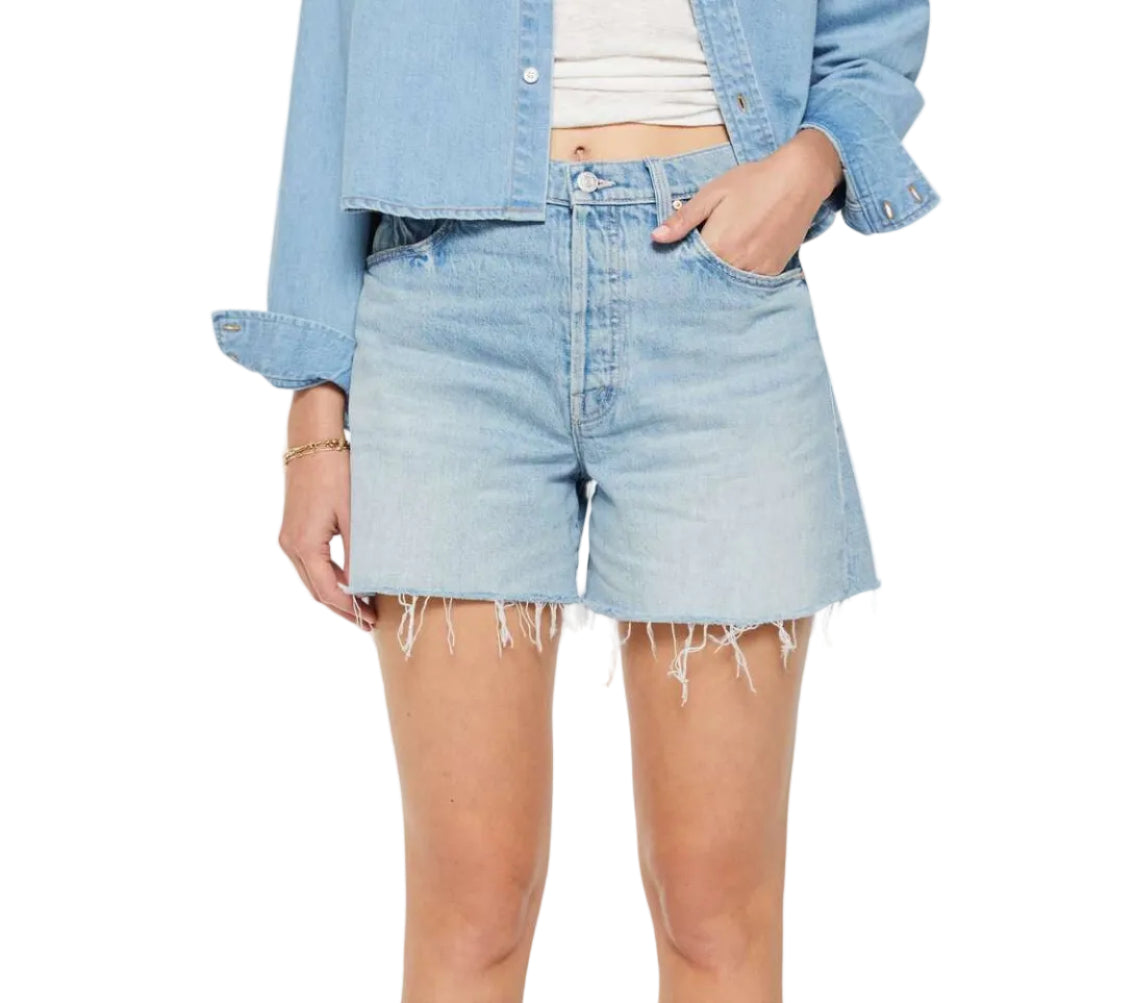 Model wearing Mother’s The Extra Mile Short Fray—high-waisted denim shorts with a frayed hem and button fly—paired with a light blue denim jacket and white top; shown from shoulders to mid-thigh with hand in pocket.