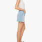 Woman standing in profile wearing a white short-sleeve top, Mother’s The Extra Mile Short Fray high-waisted denim shorts with a button fly and frayed hem, and black slide sandals against a plain white background.