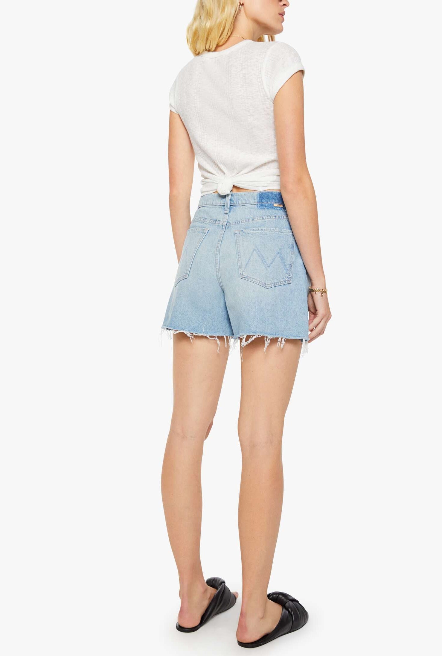 A woman stands facing away against a plain white background, wearing a white short-sleeve top and the Mother "The Extra Mile Short Fray" high-waisted denim shorts with a frayed hem, paired with black slide sandals.