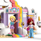 Three mini-dolls stand outside the pastel-colored LEGO® Friends Unicorn Dream Café by Legos - Toyhouse, featuring cloud and rainbow details; one is holding a unicorn-themed balloon.