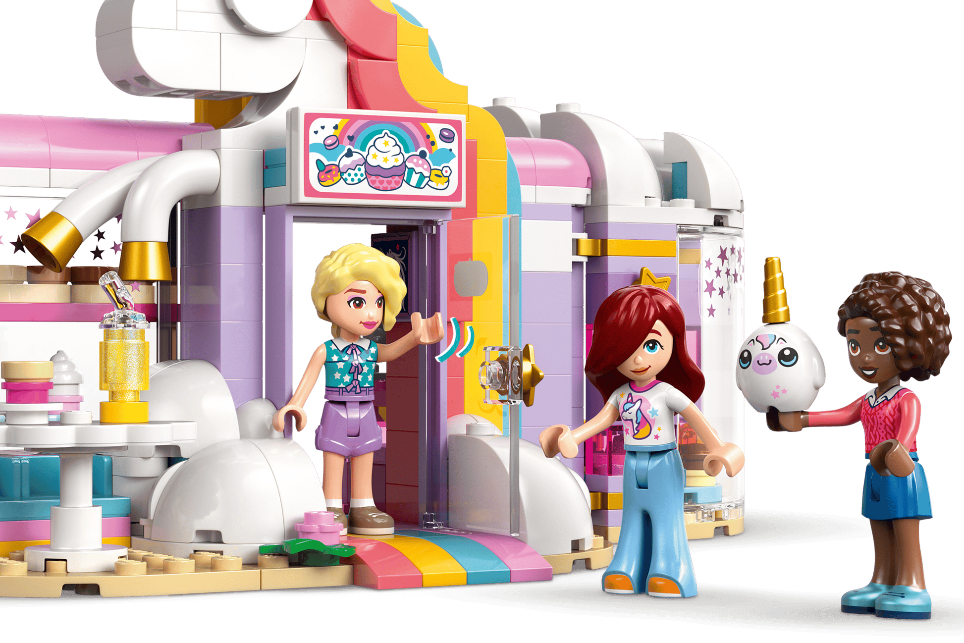 Three mini-dolls stand outside the pastel-colored LEGO® Friends Unicorn Dream Café by Legos - Toyhouse, featuring cloud and rainbow details; one is holding a unicorn-themed balloon.
