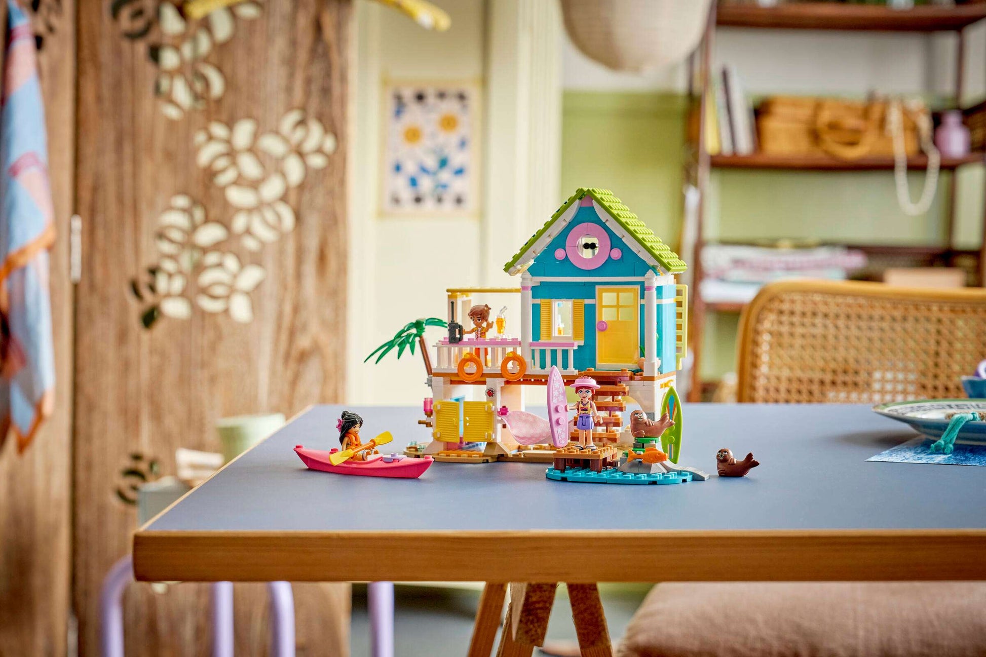 The LEGO® Friends Beach House with Seals by Legos - Toyhouse is shown with figures, a kayak, and animal seals on a table in a cozy, sunlit room featuring natural decor.