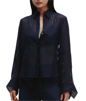 A woman wears the TWP Boyfriend Silk Organza Shirt, featuring sheer striped fabric, long sleeves with wide cuffs, and a relaxed fit, paired with dark pants.