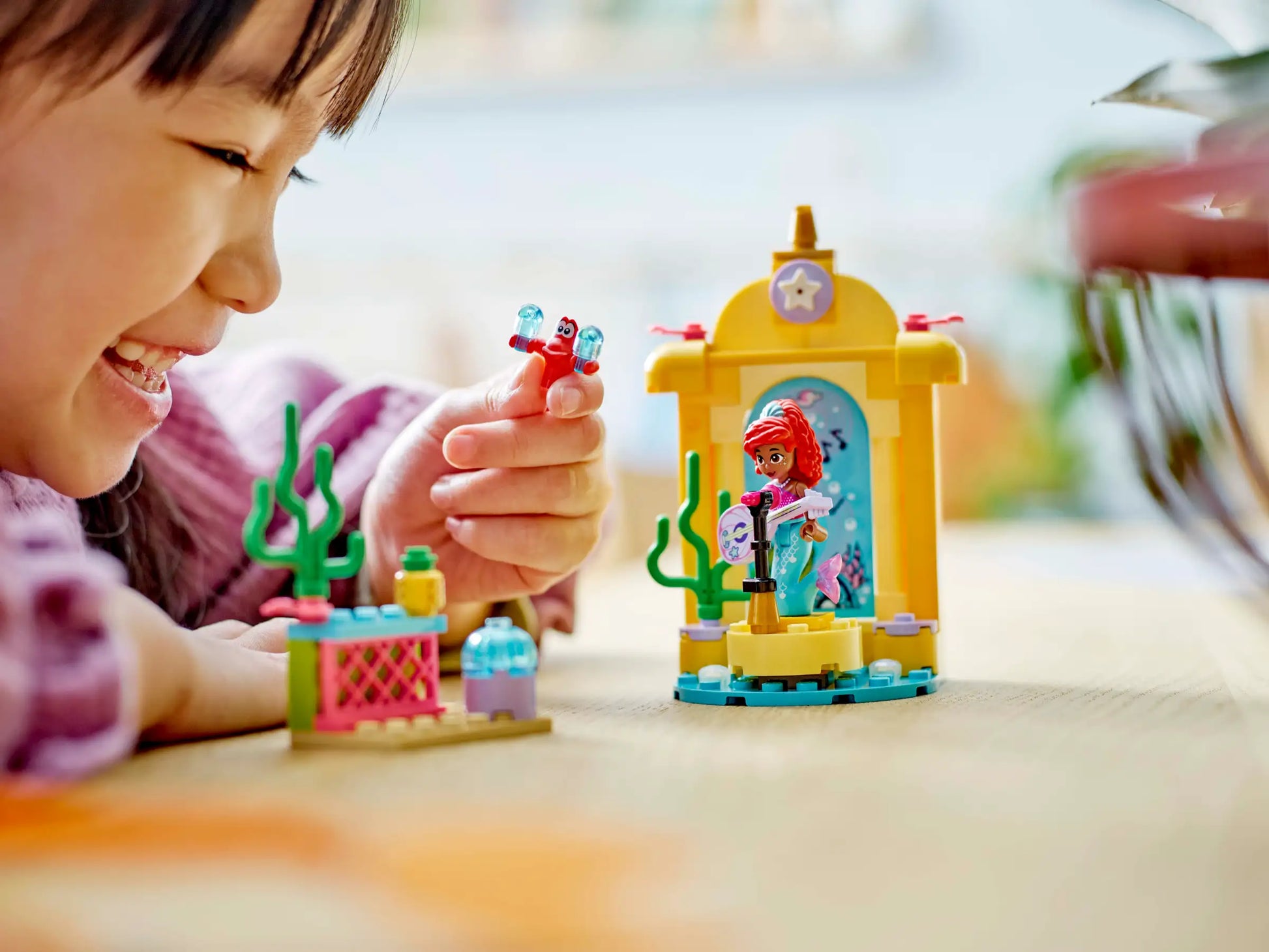 A child beams while playing with the vibrant LEGO® Disney Ariel's Music Stage set on the table, featuring beloved Disney Ariel figures and a radiant yellow structure topped with a star. This enchanting creation from Legos - Toyhouse sparks creativity and endless fun.