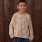 A young boy with short brown hair wears the Rylee & Cru Baby Boys’ and Boys’ Camden Long Sleeve Tee in beige and pink stripes, paired with blue jeans, as he stands in front of a wooden wall with his hands in his pockets.