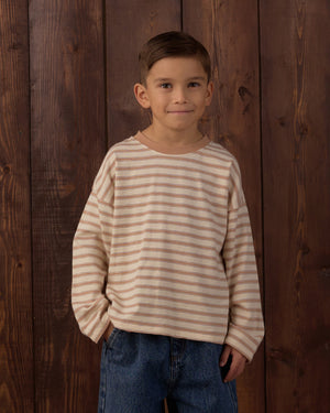 A young boy with short brown hair wears the Rylee & Cru Baby Boys’ and Boys’ Camden Long Sleeve Tee in beige and pink stripes, paired with blue jeans, as he stands in front of a wooden wall with his hands in his pockets.