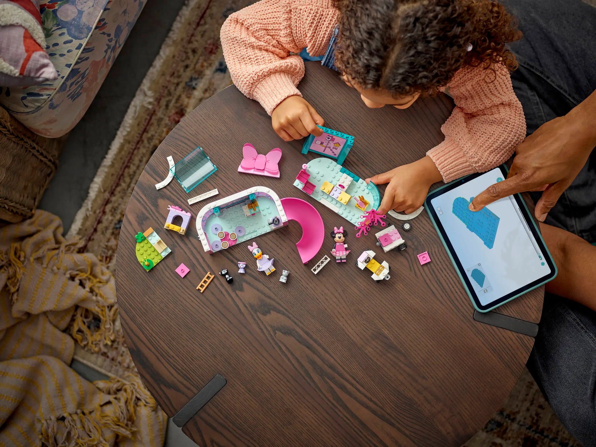 A child builds the LEGO® Disney™ Minnie's Pet Hotel from Legos - Toyhouse on a wooden table as an adult points to instructions on a nearby tablet.