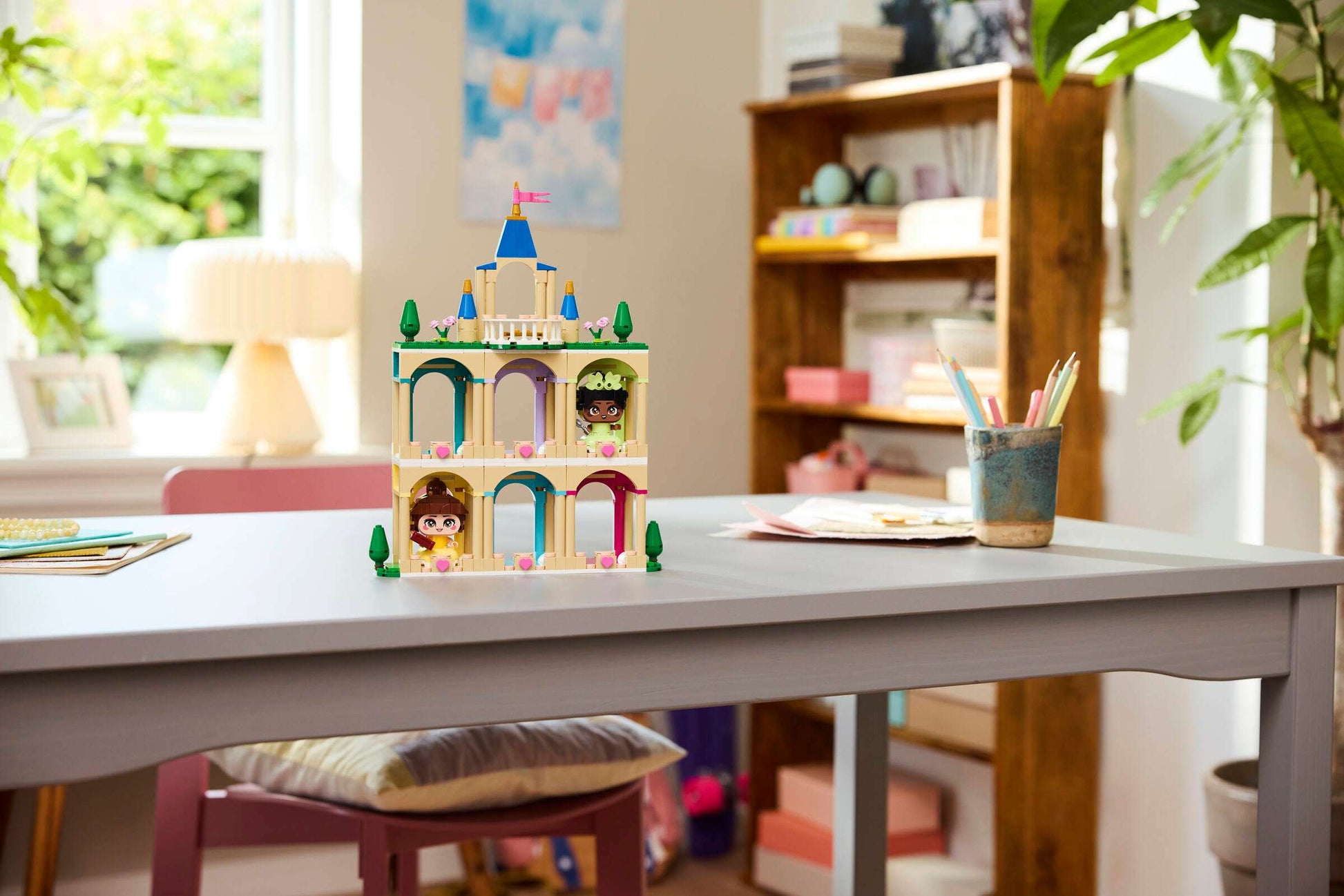 The LEGO® Disney™ Mini Belle & Tiana with Castle by Legos - Toyhouse is displayed on a gray table in a bright, tidy room with plants, shelves, and stationery.