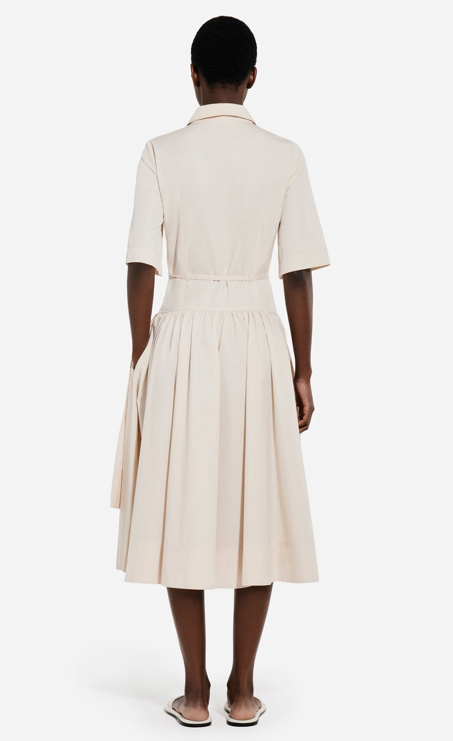 A person faces away, wearing the CO Short Sleeve Belted Cotton Poplin Dress with a collared neckline and pleated skirt—styled with flat sandals for an effortless summer essentials look.