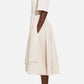 A person stands in profile wearing the CO Short Sleeve Belted Cotton Poplin Dress with a pleated skirt and pockets, paired with white flat sandals—an effortless summer essential.