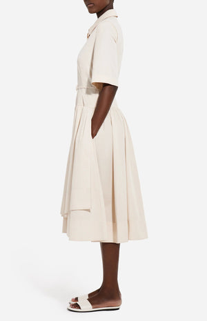 A person stands in profile wearing the CO Short Sleeve Belted Cotton Poplin Dress with a pleated skirt and pockets, paired with white flat sandals—an effortless summer essential.