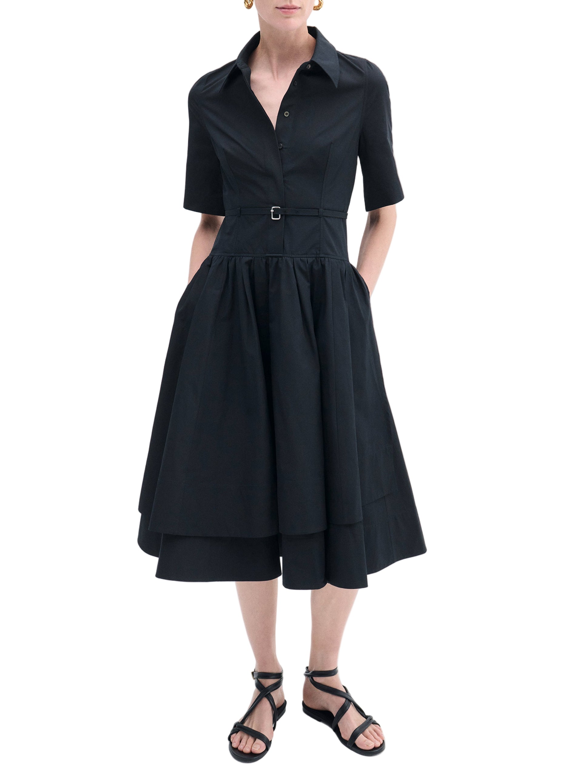 A person in the CO Short Sleeve Belted Dress—black cotton poplin, short sleeves, belted waist, midi hem—pairs it with black sandals and stands with hands in pockets.