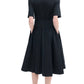 A person faces away, wearing the CO Short Sleeve Belted Dress in black cotton poplin with short sleeves, a flared midi skirt, and black sandals. The elegant dress stands out against the white background.