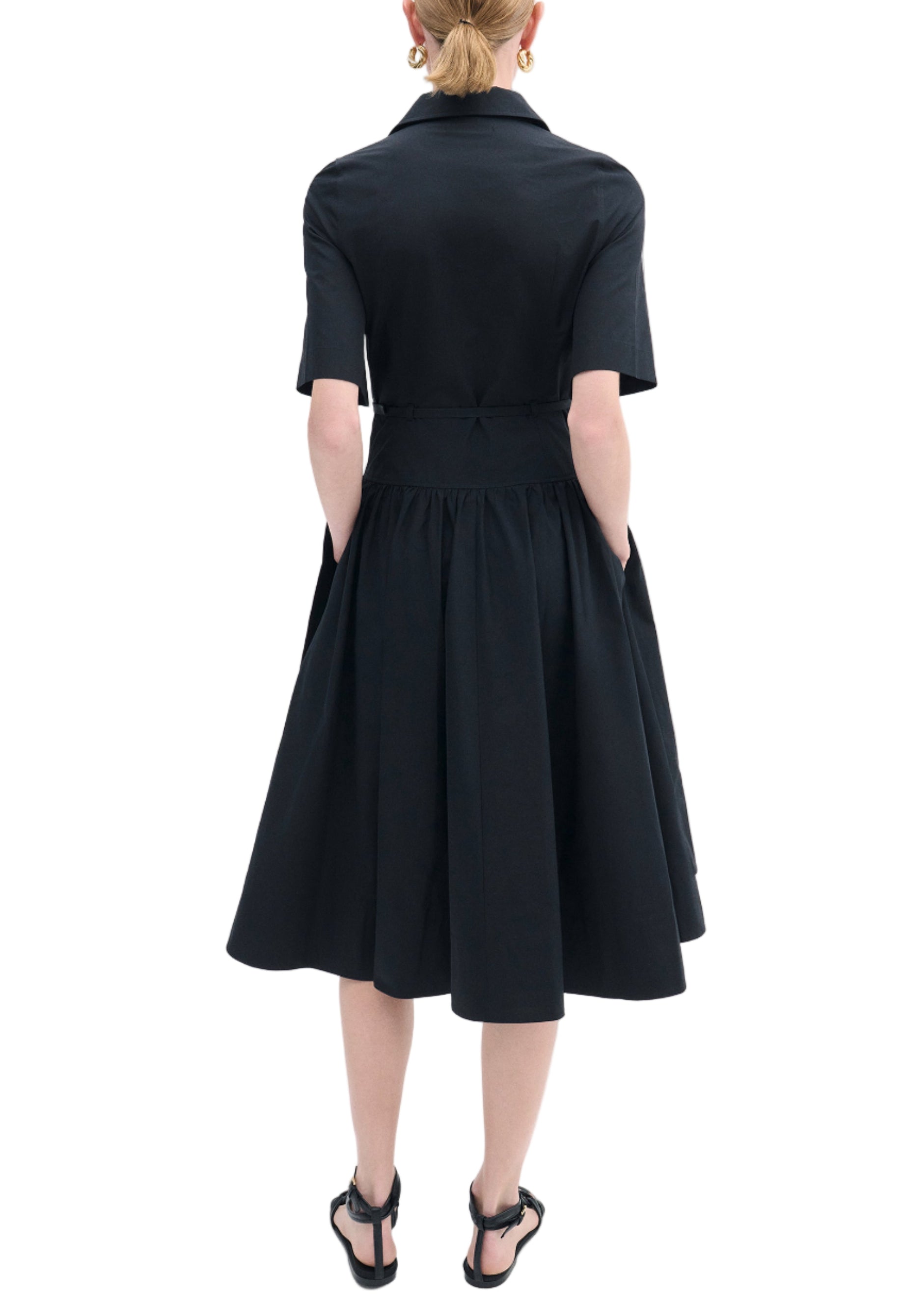 A person faces away, wearing the CO Short Sleeve Belted Dress in black cotton poplin with short sleeves, a flared midi skirt, and black sandals. The elegant dress stands out against the white background.