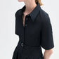 A woman wears the CO Short Sleeve Belted Dress—a black cotton poplin, midi-length shirt dress with short sleeves and a waist belt—paired with gold hoop earrings.