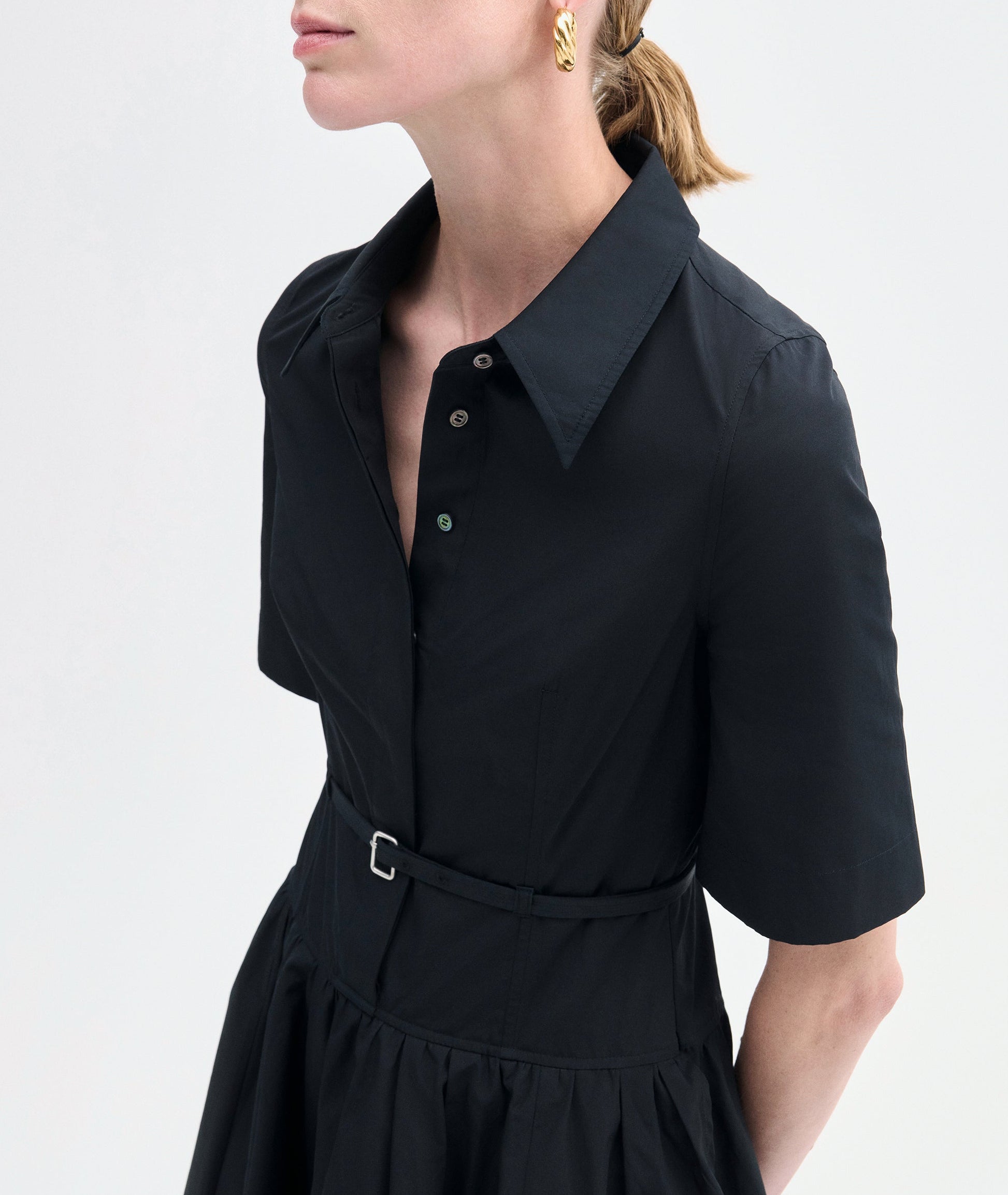 A woman wears the CO Short Sleeve Belted Dress—a black cotton poplin, midi-length shirt dress with short sleeves and a waist belt—paired with gold hoop earrings.