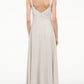A woman with long blonde hair models the Staud Madison Linen Dress, a sleeveless light beige maxi with a deep V-back, standing against a plain white background.