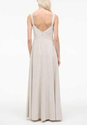 A woman with long blonde hair models the Staud Madison Linen Dress, a sleeveless light beige maxi with a deep V-back, standing against a plain white background.