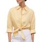 A woman wears the Staud Lisa Linen Shirt—light yellow, cropped, with a center front tie and rolled-up sleeves—paired with white high-waisted pants.