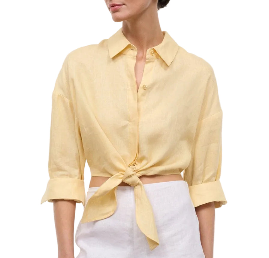 A woman wears the Staud Lisa Linen Shirt—light yellow, cropped, with a center front tie and rolled-up sleeves—paired with white high-waisted pants.