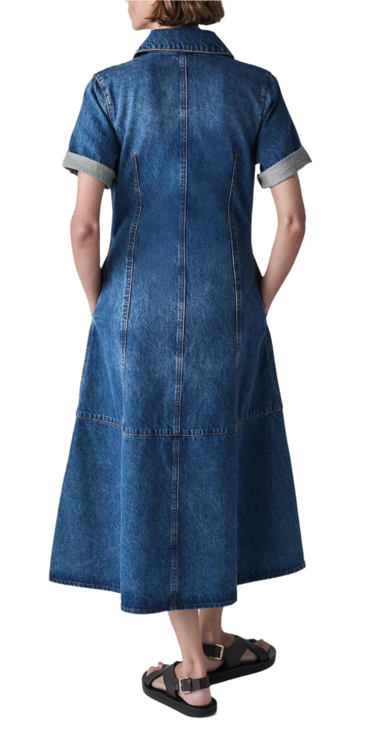 A person stands facing away, wearing the latest version of the CO Denim Midi Dress by CO, featuring rolled-up short sleeves and crafted from 100% cotton, paired with black sandals.