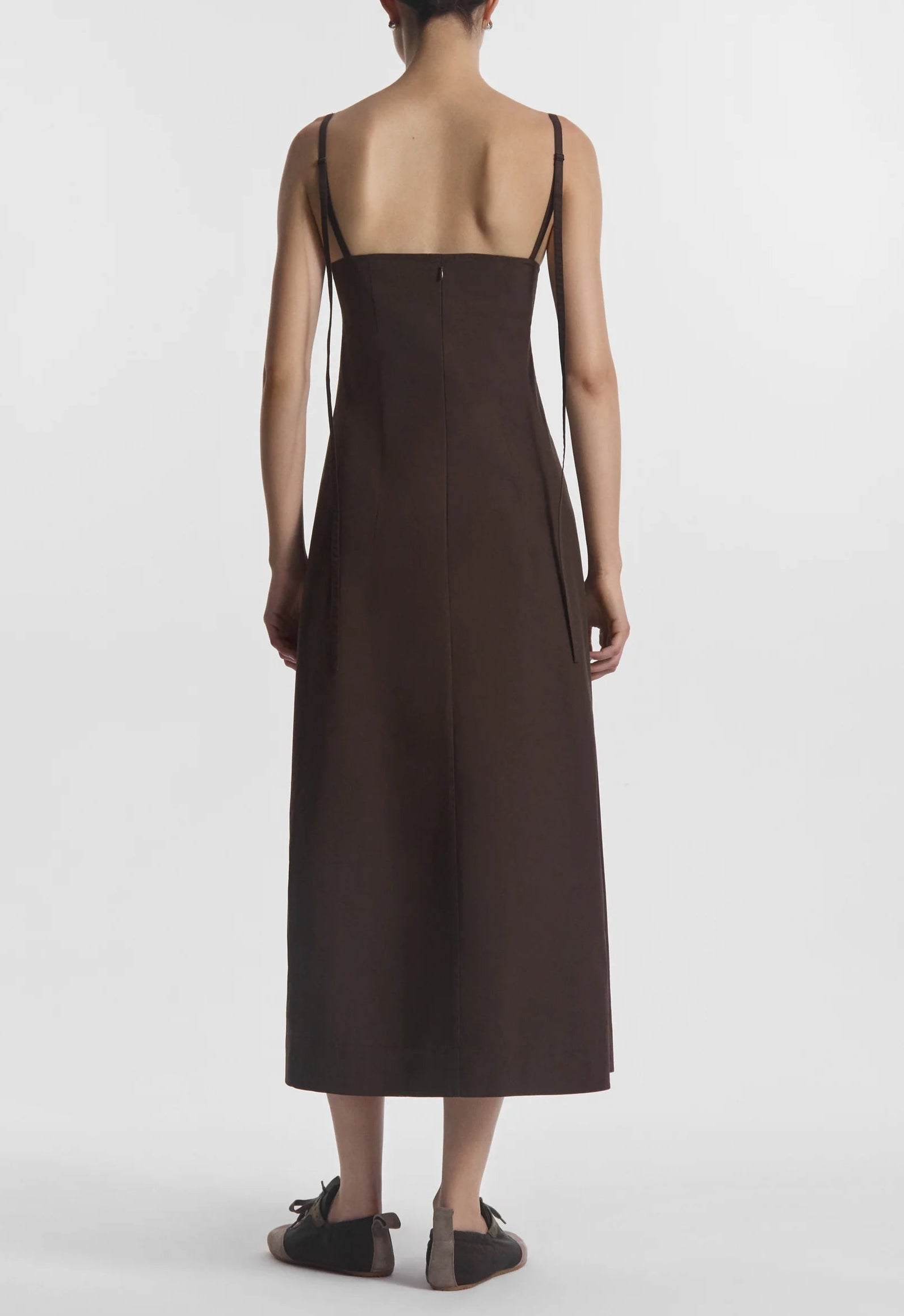 A person faces away, wearing the CO Bustier Cut Dress—a sleeveless dark brown cotton poplin dress with thin straps—paired with flat shoes, set against a plain white background.