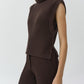A woman wearing the Maple Top, a sleeveless brown ribbed turtleneck sweater by Simkhai, paired with brown pants, stands against a plain light background.