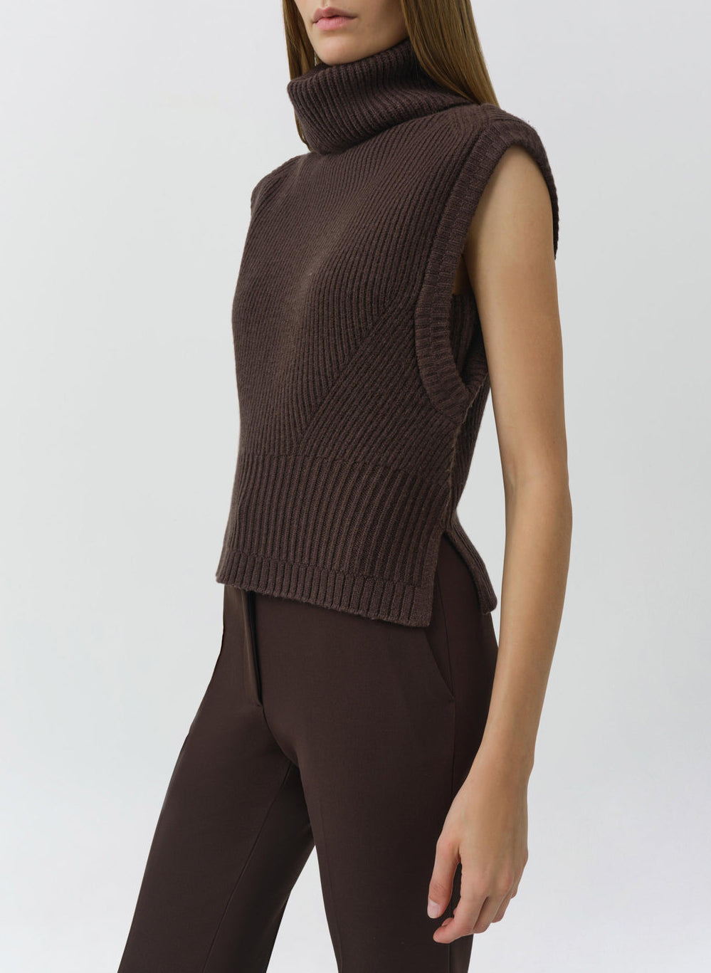 A woman wearing the Maple Top, a sleeveless brown ribbed turtleneck sweater by Simkhai, paired with brown pants, stands against a plain light background.